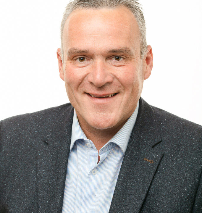 Homme souriant en costume sombre et chemise bleue, regardant directement l&rsquo;appareil photo.