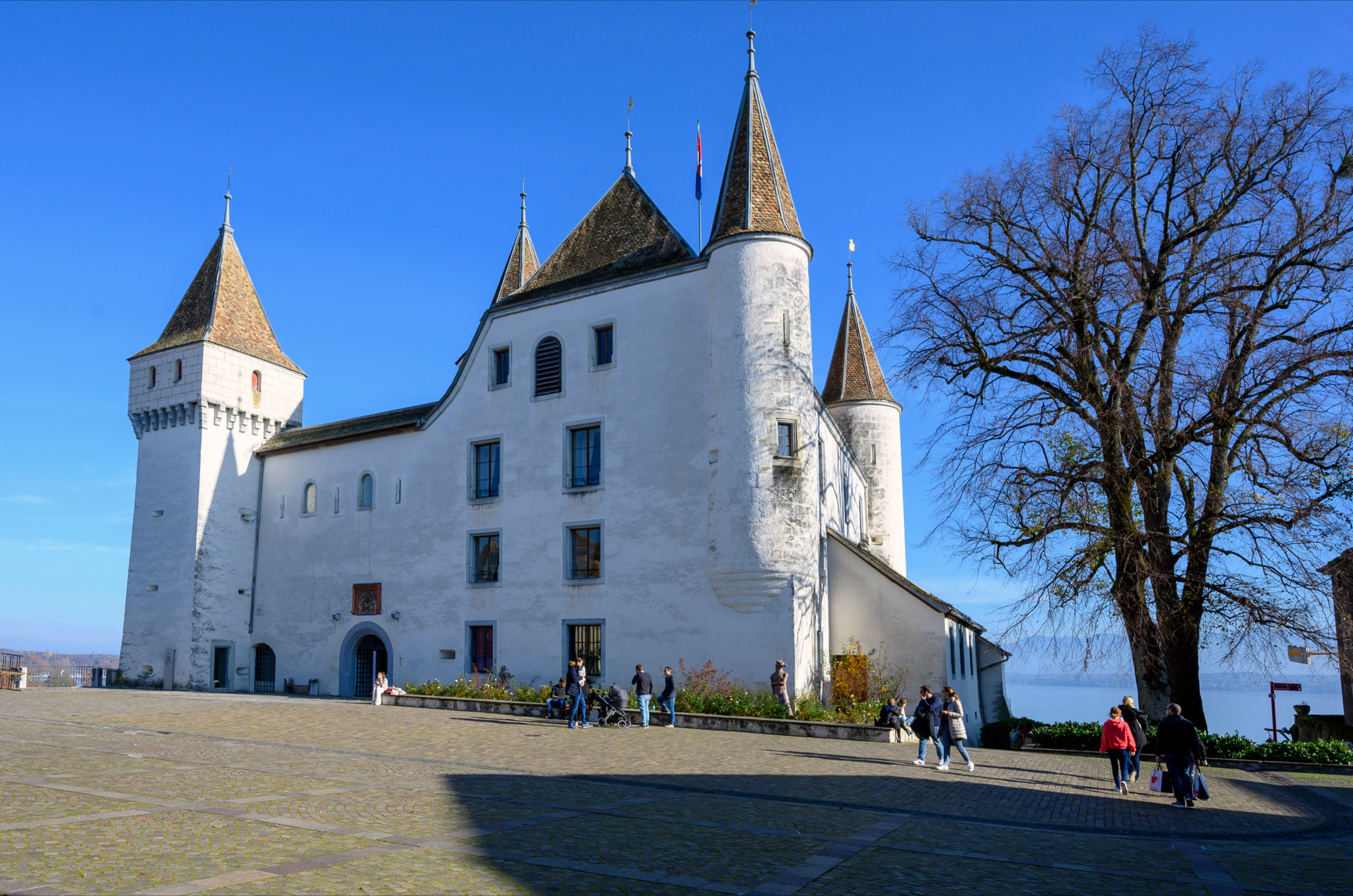 Le Ch&acirc;teau de Nyon avec ses tours historiques et des visiteurs se promenant sur la place, sous un ciel bleu, novembre 2020.
