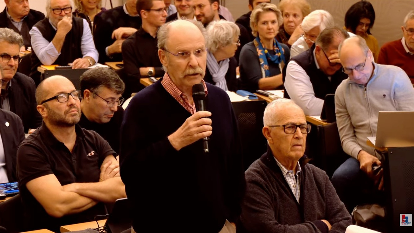 Un homme en pull noir parle dans un microphone lors d&rsquo;une conf&eacute;rence avec un public attentif assis derri&egrave;re lui.
